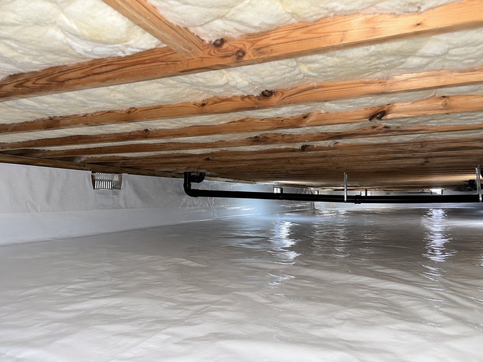 Spray foam insulation filling the bays between floor joists on the underside of a subfloor, above a crawl space lined with white plastic sheeting, with a black drain pipe running along the wall