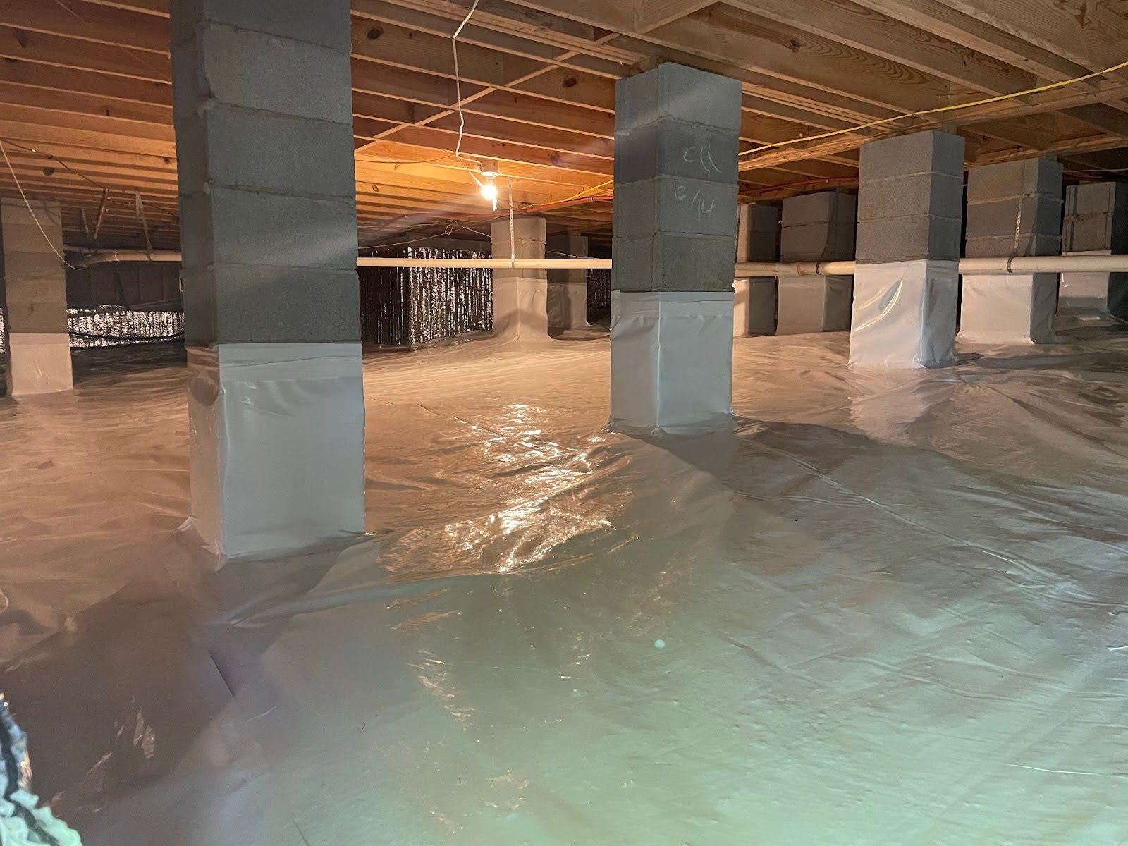 A Portland crawl space mid-encapsulation: white reinforced vapor barrier across the soil, green closed-cell spray foam sealing the rim joists, red plumbing supply lines overhead
